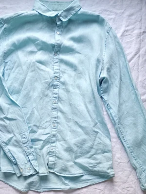Ljusblå skjorta i bomull - Säljer en stilren turkos skjorta med klassisk krage och knappar framtill. Skjortan är långärmad och tillverkad i linne, perfekt för en clean och enkel look.