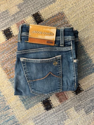 Limited edition selvage Jacob Cohen - Säljer ett par klassiska blå jeans från Jacob Cohen med raka ben och snygga slitningar framtill. Jeansen har fem fickor, kontrastsömmar och en läderpatch med logga bak i midjan. Perfekta för en avslappnad och stilren look.