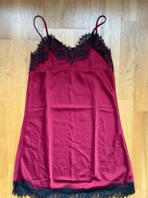 Vinrött nattlinne med spetsdetaljer - Snyggt vinrött nattlinne med tunna axelband och svarta spetsdetaljer vid byst och nederkant. Materialet är glansigt och känns mjukt mot huden, perfekt för en chill kväll hemma. Spetsen ger en extra lyxig känsla och nattlinnet har en feminin siluett.