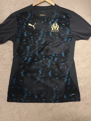 Marseille träningströja Puma svart/blå - Snygg svart träningströja från Puma med Olympique de Marseille-logga i gult på bröstet. Tröjan har blått mönster framtill, rund hals och korta ärmar. Tillverkad i funktionsmaterial DryCell som andas bra. Perfekt för fotbollsträning eller gym.