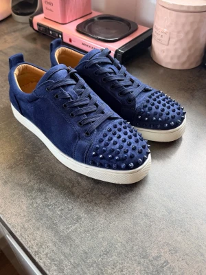 Blåa sneakers med nitar från Louboutin - Snygga mörkblå sneakers i mocka från Christian Louboutin med coola matchande nitar på tån och sidorna. Skorna har vita sulor och den klassiska röda undersidan. Perfekta för dig som vill sticka ut med din stil. Köpte den i höstas och har använt varsamt. På sista bilden syns det ett en nit fattas, det är den enda skadan på skorna. Andra nitar har limmats fast men de sitter på, skriv vid några frågor