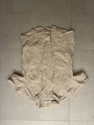 Beige linneskjorta från H&M - Säljer en beige skjorta från H&M i luftigt linne. Skjortan har korta ärmar, klassisk krage och knappar framtill. Perfekt för varma dagar och enkel att styla till olika outfits.
