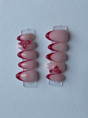 Press-on naglar med röd fransk tipp - Snygga press-on naglar i mandelform med ljusrosa bas och röd fransk tipp. Två av naglarna har dekorativa blommor med små pärlor för extra detalj. Perfekt för dig som vill ha en trendig och unik look.