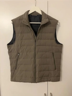 Beige väst - Stilren beige dunväst med quiltat mönster och hög krage. Västen har två framfickor med dragkedja och stängs med en hel dragkedja framtill. Perfekt för lager-på-lager och ger en clean look till din outfit.