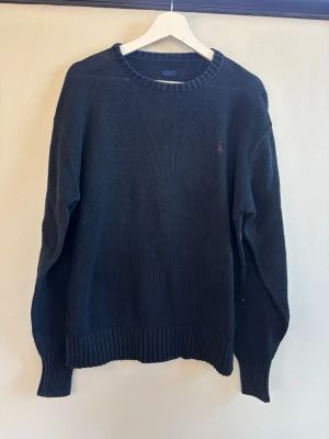 Mörkblå stickad tröja från Polo Ralph Lauren - Mörkblå stickad tröja från Polo Ralph Lauren med rund halsringning och diskret röd logga broderad på bröstet. Tröjan har ribbstickade muddar vid ärmslut och nederkant. Perfekt för lager-på-lager och en klassisk look.