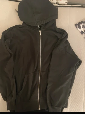 Svart zip hoodie med huva - En svart hoodie med dragkedja framtill och stor huva. Klassisk modell med två fickor på magen och ribbade muddar vid ärmslut och nederkant. Perfekt för en chill och avslappnad stil. Materialet känns mjukt och bekvämt, troligen bomull eller bomullsblandning.