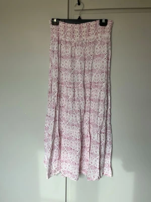 Långkjol med rosa mönster - Långkjol i vitt med ljusrosa och mörkrosa blommigt mönster. Kjolen har en luftig och flowy passform med resår i midjan. 93cm lång 