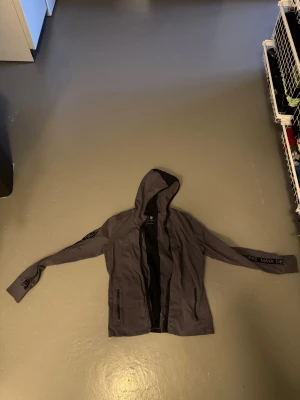 Grå hoodie från Maya Delorez - Snygg grå hoodie från Maya Delorez med dragkedja framtill och svarta detaljer. Huva och två fickor med dragkedja. Svart text 'MAYA DELOREZ' längs ärmarna ger en sportig vibe. Perfekt till stallet för kyliga dagar. Stl L. Kom gärna med bud💗