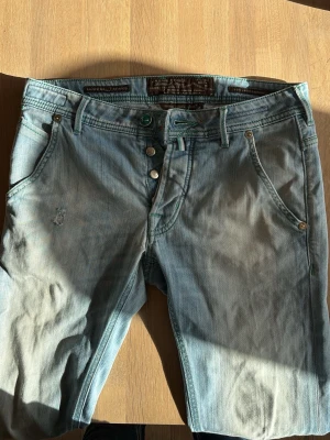 Jacob Cohen jeans - Säljer ett par ljusblå jeans från Jacob Cohen med klassisk femficksmodell och knappgylf. Jeansen har en snygg tvättad look, gröna sömmar och patch med logga bak. Tillverkade i mjuk denim med en avslappnad passform.