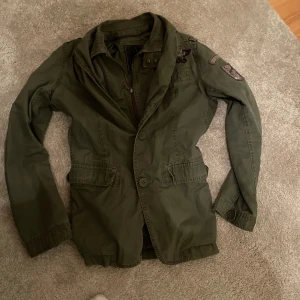 Field Jacket - Snygg militärgrön Field Jacket med coola patchar på axeln och armen. Jackan har både dragkedja och knappar framtill, samt två fickor med lock. Passformen är normal och materialet känns som bomull. Perfekt för dig som gillar en avslappnad men stilren look.