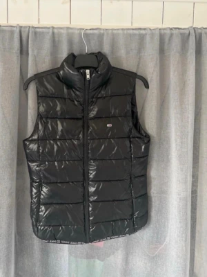 Svart dunväst från Tommy Jeans - Stilren svart dunväst från Tommy Jeans med quiltad design och hög krage. Västen har en diskret logotyp på bröstet och dragkedja framtill. Materialet är glansigt och lätt, perfekt för lager-på-lager och att hålla värmen under kyliga dagar.