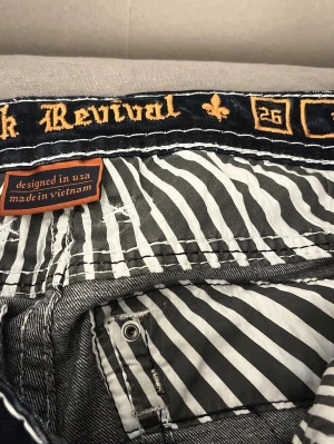 Rock Revival bootcut jeans.  - Snygga blå bootcut/baggy jeans från Rock Revival med coola broderade detaljer på bakfickorna och kontrastsömmar. Jeansen har dragkedja, knappar och insida med svartvita ränder. Jeansen nuddar nästan marken på mig som är runt 170cm. Jeansen är oanvända, köpta på nätet. dom är ganska stretchiga. Skulle passa storlek w26, w28💕Orgenal pris 2000kr