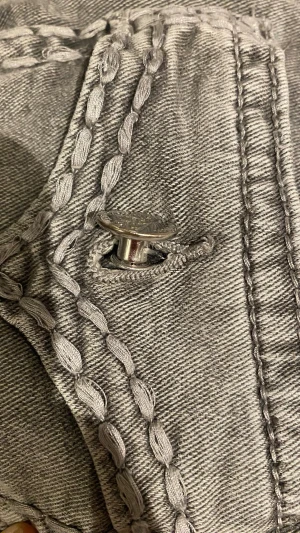 Grå jeans med dekorativa sömmar - Säljer ett par grå jeans med unika, dekorativa sömmar längs fickor och ben. Jeansen har en klassisk femficksmodell, knappgylf och smal passform. Materialet är denim för extra komfort. Perfekta för dig som vill sticka ut lite extra!