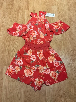 Röd blommig off shoulder jumpsuit - Superfin röd jumpsuit med stora blommor i rosa och orange toner. Off shoulder-modell med volangärmar och smockad midja för en snygg passform. Lätt och luftig, perfekt för sommardagar. Halterneck med liten öppning framtill ger en trendig look.