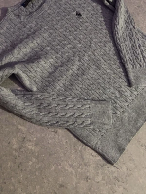 Grå kabelstickad tröja Polo Ralph Lauren - Kabelstickad tröja i ljusgrått från Polo Ralph Lauren med rund hals och ribbade muddar. Tröjan har en liten broderad logga på bröstet och är gjord i mjukt material. Perfekt för dig som gillar klassisk och stilren look.