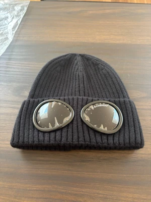 Cp company mössa svart - Säljer en svart ribbstickad mössa från C.P. Company med ikoniska inbyggda glasögon på uppviket. Mössan är tillverkad i mjukt material och har en klassisk beanie-form som sitter snyggt på huvudet. Perfekt för dig som vill sticka ut med en streetwear-inspirerad stil.