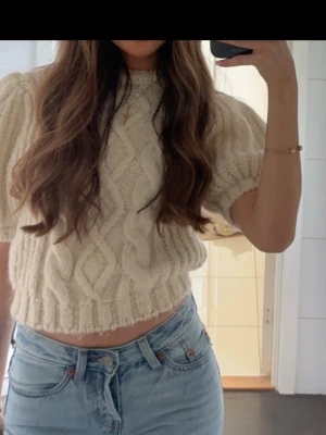 Vit kabelstickad tröja från Zara - Trendig vit kabelstickad tröja från Zara med puffärmar och kortare, boxig passform. Tröjan har grovstickat mönster och ribbade muddar vid ärmslut och nederkant. Perfekt till jeans för en avslappnad men snygg look.