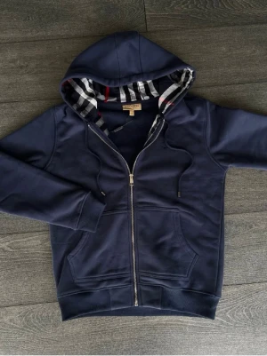 Marinblå hoodie från Burberry - Snygg marinblå hoodie från Burberry med dragkedja och klassiskt rutigt foder i huvan. Hoodien har fickor framtill, ribbade muddar och justerbar dragsko i huvan. Perfekt för dig som gillar stilrena detaljer och exklusiv känsla.