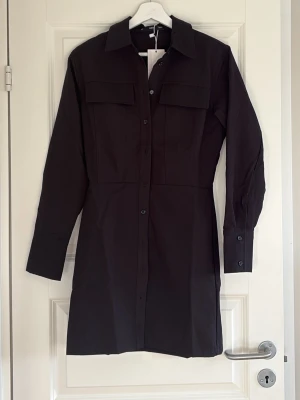 Svart skjortklänning från Mango - Stilren svart skjortklänning från Mango med knappar framtill, två bröstfickor och klassisk krage. Klänningen har lång ärm med manschett och är i en rak, normal passform. Perfekt att styla med boots eller sneakers.