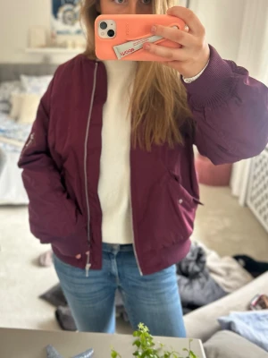 Vinröd bomberjacka med dragkedja - Snygg vinröd bomberjacka med silverfärgad dragkedja och ribbade muddar. Jackan har två fickor med knapp framtill och klassisk bomberpassform. Perfekt för dig som vill ha en stilren och trendig look. Passar dig som vill ha en oversize look i ca S❤️