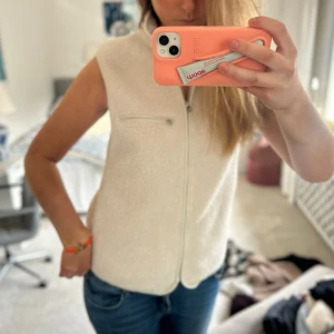 Vit fleeceväst med dragkedja - Säljer en krämvit fleeceväst med hög krage och dragkedja framtill. Västen har en ficka med dragkedja på bröstet och är ärmlös, perfekt att slänga över en hoodie eller tröja för extra värme. Enkel och clean stil som passar till det mesta. Passar ca M🥳