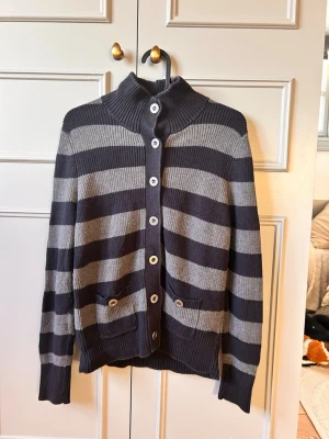 Randig kofta från Tommy Hilfiger - Snygg kofta från Tommy Hilfiger i mörkgrått och ljusgrått med breda ränder. Koftan har hög krage, knäppning med vita knappar framtill och två fickor med knappdetaljer. Tillverkad i mjukt stickat material, perfekt för lager på lager. Stl (M) allt är sprillans ny