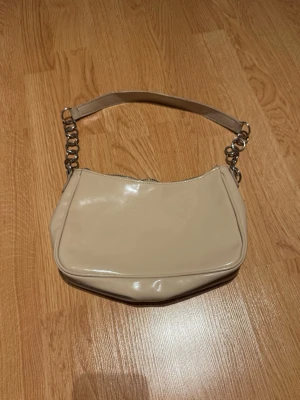Beige axelväska med kedjedetaljer - Snygg beige axelväska i glansigt material med silvriga kedjedetaljer på axelremmen. Bandet har några repor! 