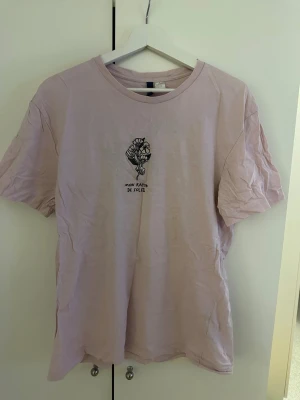 Ljusrosa t-shirt med tryck - Ljusrosa t-shirt med svart tryck av blommor och texten 'Mon rayon de soleil' på bröstet. På ryggen finns ett stort fototryck med rosa blommor och blå himmel. T-shirten har rund hals och korta ärmar, perfekt för en chill och stilren look.