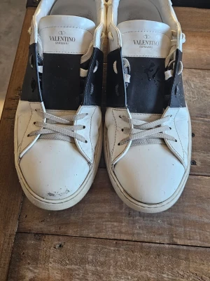 Valentino Open Garavani sneakers (äkthetsbevis) - Säljer ett par Valentino Open Garavani sneakers i vitt och svart skinn med ikoniska nitar runt hälen. Skorna har snörning och platt sula, med en chunky och modern siluett. Medföljer originalkartong, dustbags och extra skosnören. Skorna är i bra skick men lite slitage vid tån på höger sko. Skorna är smutsiga men går att tvätta bort enkelt med ljummet vatten. Pris kan diskuteras! Hör av er om intresserad!