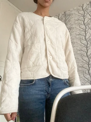 Vit quiltad cardigan med knappar - Stilren vit cardiganjacka med quiltat mönster och knappar framtill. Jackan har en rak passform, rund halsringning och långa ärmar. Perfekt att slänga över en t-shirt för en avslappnad look.