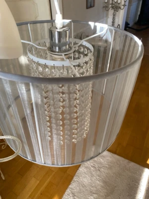 Ny taklampa från Homeroom - Säljer en ny, oanvänd  taklampa med ljusgrå lampskärm i tyg och dekorativa hängande prismor i mitten. Lampan har en modern känsla med metallfäste upptill och transparent sladd. Perfekt för att ge rummet en lyxig touch. Nypris 1399kr. Mått mm se bilderna.Finns i Bandhagen