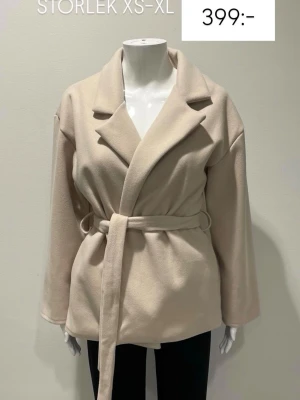 Beige kappa med skärp från missäy  - Stilren beige kappa med bred krage och knytskärp i midjan. Kappan har raka ärmar och är tillverkad i ett mjukt material som ger en avslappnad och modern look. Perfekt för att skapa en trendig outfit under våren