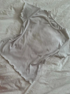 Vit ribbad topp med vågig kant - Säljer en vit ribbad topp med korta ärmar och vågiga kanter vid ärmslut och nederkant. Toppen har en rund halsringning och är i ett mjukt, stretchigt material som sitter snyggt på kroppen. Perfekt att styla med jeans eller kjol.