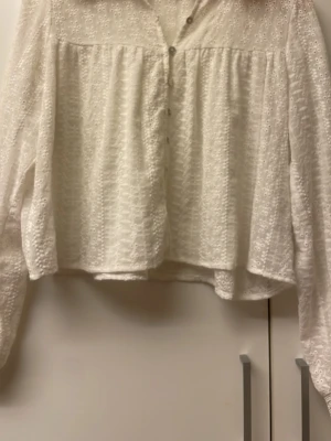 Vit broderad blus med knappar - Superfin vit blus med broderade blommor och knappar framtill. Blusen har långärmade ärmar med breda spetsmuddar och är gjord i ett tunt, luftigt tyg med struktur. Perfekt att styla med jeans eller kjol för en trendig look.