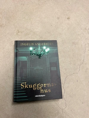 Skuggornas hus av Ingelin Angerborn - En mystisk och spännande ungdomsroman av Ingelin Angerborn. Boken har ett svart och grönt omslag med en gammal trappa och en ljuskrona, tryckt i hårdpärm. Perfekt för dig som gillar mysterier och övernaturliga berättelser.