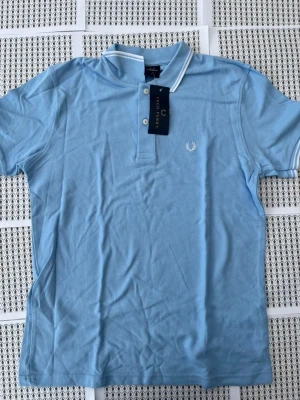 Ljusblå pikétröja från Fred Perry - Snygg ljusblå pikétröja från Fred Perry med klassisk krage och två knappar framtill. Tröjan har korta ärmar och vit logga broderad på bröstet. Tillverkad i mjuk bomull och har vita detaljer på krage och ärmslut.