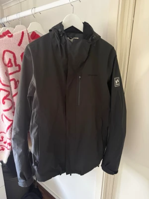 Svartgrå vindjacka från 24 - Snygg svartgrå vindjacka från 24 med huva och dragkedja. Jackan har en bröstficka med dragkedja och diskret logga på bröstet samt ett märke på ärmen. Tillverkad i ett lätt och vindtätt material, perfekt för blåsiga dagar.