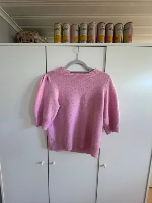 Ljusrosa stickad tröja Vero Moda - Supersöt ljusrosa stickad tröja från Vero Moda med puffärmar och ribbade muddar. Tröjan har rund halsringning och är gjord i ett mjukt material som känns skönt mot huden. Perfekt för dig som gillar en feminin och trendig look.