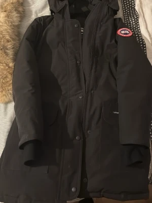 Svart trillium parka -  Canada Goose -  Canada Goose med huva i modellen trillium parka,  avtagbar päls. Jackan har två stora framfickor, dragkedja och tryckknappar framtill samt den klassiska loggan på ärmen. Perfekt för kalla vinterdagar och riktigt varm. Jackan är i toppskick då den endast varit använd cirka 3 gånger, köpt från miinto, kvitto finns!