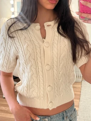 Beige stickad kofta från H&M - Kortärmad kabelstickad kofta i vit färg från H&M. Koftan har stora matchande knappar framtill, ribbade kanter och en croppad passform. Perfekt att styla med jeans eller kjol för en trendig look.