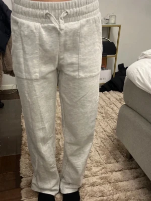 Ljusgrå mjukisbyxor med dragsko - Säljer ett par ljusgrå mjukisbyxor med bred resår i midjan och dragsko. Byxorna har raka ben och stora fickor framtill. Tillverkade i mjukt och bekvämt material, perfekta för chill dagar. Passar dig som gillar en avslappnad stil.