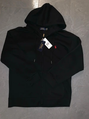 Svart hoodie från Polo Ralph Lauren - Snygg svart hoodie från Polo Ralph Lauren med dragkedja framtill och klassisk huva. Märkets ikoniska röda logga är broderad på bröstet. Hoodien har dragsko i huvan och är tillverkad i mjukt bomullsmaterial. Perfekt för en avslappnad och stilren look.