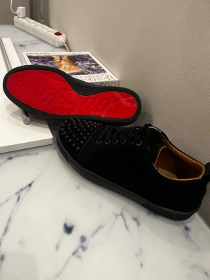 Svarta Louboutin sneakers med nitar - Säljer ett par svarta sneakers från Christian Louboutin med klassisk röd sula och svarta nitar på tån. Skorna har snörning, är låga och har en lyxig känsla med mockaliknande material och läderdetaljer på insidan.