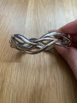 Silvrigt flätat armband - Snyggt armband i silverfärgad metall med flätad design. Armbandet har en stilren och modern look med mjuka, böljande former. Perfekt för dig som gillar minimalistiska accessoarer med en twist. Lätt att matcha med olika outfits och stängs med ett smidigt spänne.