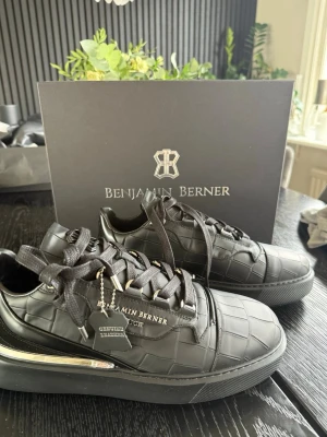 Svarta sneakers från Benjamin Berner - Stilrena svarta sneakers från Benjamin Berner med croco-mönstrad läderlook och svarta skosnören. Snygga metalldetaljer på sidan och logotyp på plösen och sidan. Perfekta för dig som gillar exklusiv och modern design.