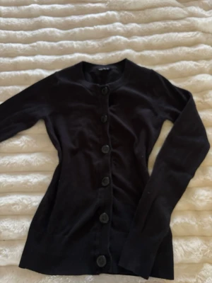 Vintage svart kofta med stora knappar - En klassisk svart kofta med rund halsringning och stora svarta knappar framtill. Långärmad och i mjukt, tunt stickat material som passar perfekt till lager-på-lager. Enkel och stilren design som funkar till det mesta. Köpte secondhand för 500kr, lappen var avklippt när jag köpte den så vet inte vart den är ifrån.