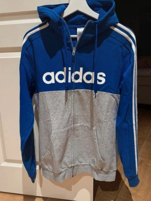 Blå/grå adidas hoodie - Snygg hoodie från adidas i blått och grått med dragkedja framtill och stor vit logga över bröstet. Klassiska vita ränder längs ärmarna och praktisk huva med dragsko. Två fickor framtill. Perfekt för en sportig och avslappnad stil.