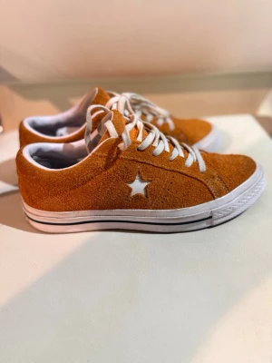 Converse One Star orange mocka sneakers - Säljer ett par Converse One Star sneakers i orange mocka med vit stjärna på sidan och vita snören. Skorna har vit sula med svart rand och klassisk låg profil. Perfekta för dig som vill sticka ut med färg och stil.