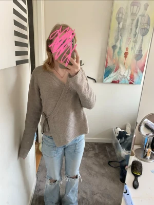 Beige omlottkofta från H&M - Säljer en beige stickad omlottkofta från Lager 157 med knytband i sidan. Koftan har v-ringning, långa ärmar och ribbade muddar. Perfekt att slänga över ett linne för en chill och snygg look.