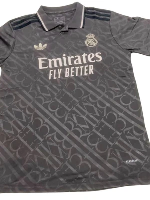 Real Madrid svart matchtröja Adidas 24/25 third  - Snygg svart Real Madrid matchtröja från Adidas med diskret mönster, klubbmärke och Adidas-logga på bröstet. Tryck med 'Emirates Fly Better' på framsidan och Aeroready-material för bra ventilation. Klassisk krage och korta ärmar.